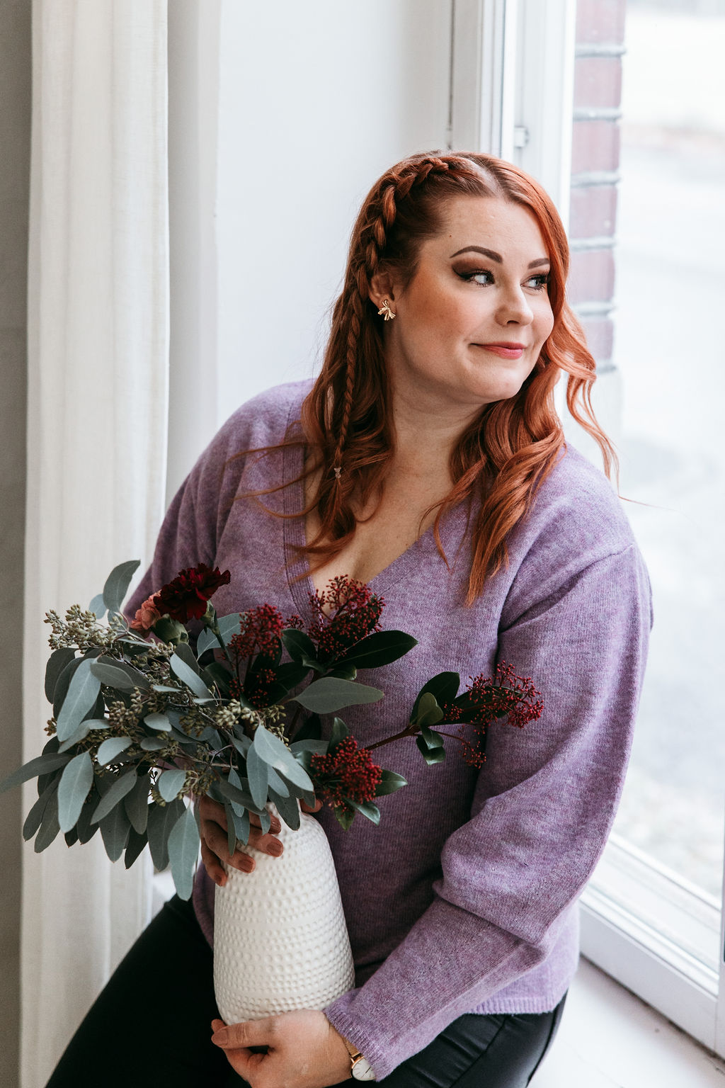 Sisustussuunnittelija Meri Rapo istuu ikkunan ääressä ja pitelee kukkavaasia, katse suunnattuna ulos.