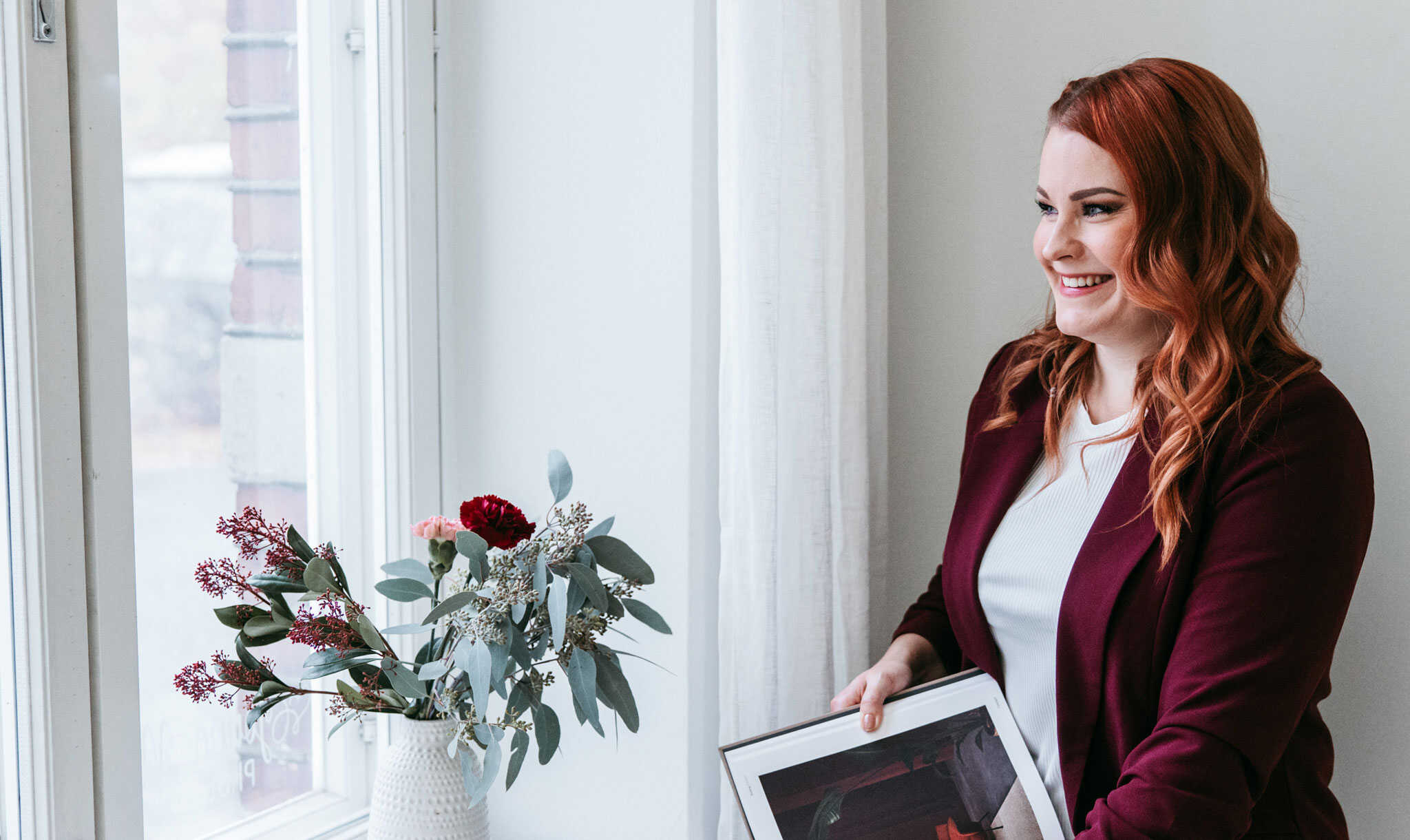 Tila- ja sisustussuunnittelija Meri Rapo istuu ikkunan ääressä hymyillen ja pitää sisustussuunnitteluun liittyvää kirjaa sylissään.